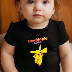 May include: A black baby onesie with the text "Shockingly Cute" in a colorful gradient. A yellow Pikachu graphic is printed below the text. The onesie has short sleeves and a round neckline.