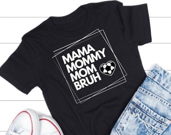 May include: Black t-shirt with white text that reads "Mama Mommy Mom  Bruh" with a soccer ball inside a heart. The shirt is laid out on a white wood surface with a pair of white sneakers and a pair of light blue denim shorts.