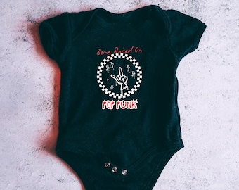 May include: Dark green baby bodysuit with the text "Being Raised On Pop Punk". The design features a white and black checkered circle with a hand gesture and musical notes. The bodysuit has three snaps at the bottom.
