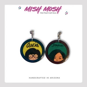 May include: A pair of earrings featuring Daria and Jane from the animated series Daria. The earrings are round and have a yellow and green background with the characters' faces and names. The earrings are handcrafted in Arizona.