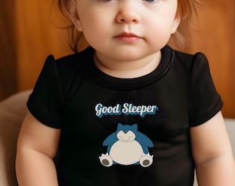 May include: A black baby onesie with the text "Good Sleeper" in a script font above a cartoon illustration of a sleeping blue creature. The baby is looking directly at the camera.