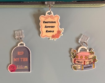 May include: Three decorative keychain charms. One reads "Emotional Support Kindle" with floral accents. Another is a tombstone with "RIP MY TBR". The third charm depicts a stack of books with "FULLY BOOKED".