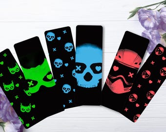 May include: A collection of black bookmarks with various designs. One features a blue skull with heart eyes, another a green devil face, and others with skulls, hearts, and crosses. The bookmarks are arranged on a white wooden surface.