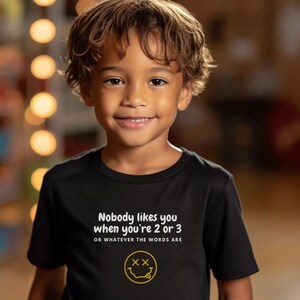 May include: A black t-shirt with white text that reads "Nobody likes you when you're 2 or 3 or whatever the words are". A yellow smiley face with an "X" for eyes is below the text.