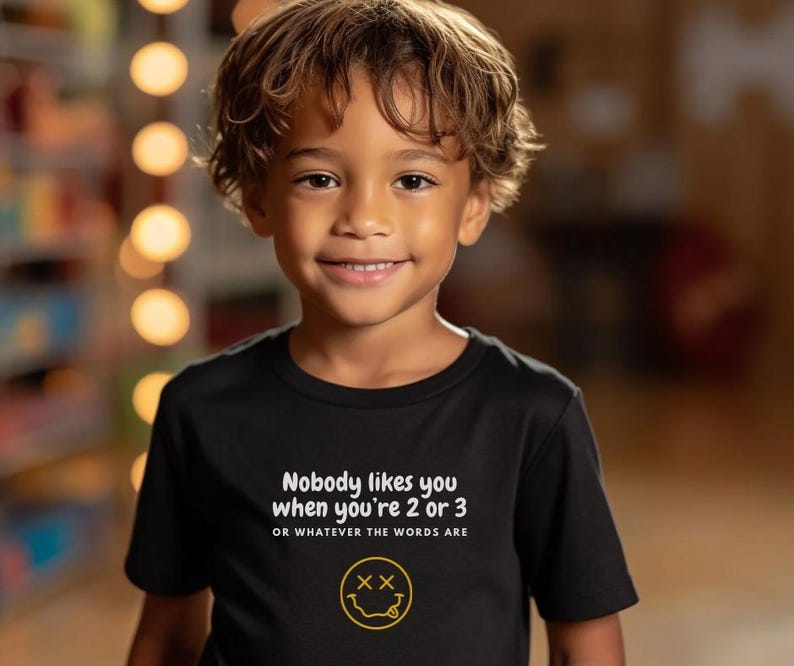 May include: A black t-shirt with white text that reads "Nobody likes you when you're 2 or 3 or whatever the words are". A yellow smiley face with an "X" for eyes is below the text.