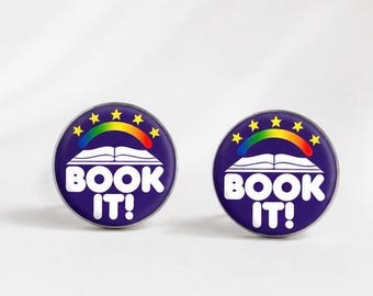 May include: Two round, purple pins with the text "BOOK IT!" in white. Each pin features a rainbow over an open book, surrounded by yellow stars. The pins are likely for book lovers.