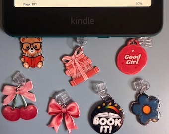 May include: A Kindle e-reader with several decorative charms attached. Charms include a bear reading a book, a gift, a red circle with "Good Girl", cherries, a pink bow, a charm with "BOOK IT!", and a blue flower.