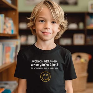 May include: A black t-shirt with white text that reads "Nobody likes you when you're 2 or 3 OR WHATEVER THE WORDS ARE" and a yellow smiley face with an X for eyes.