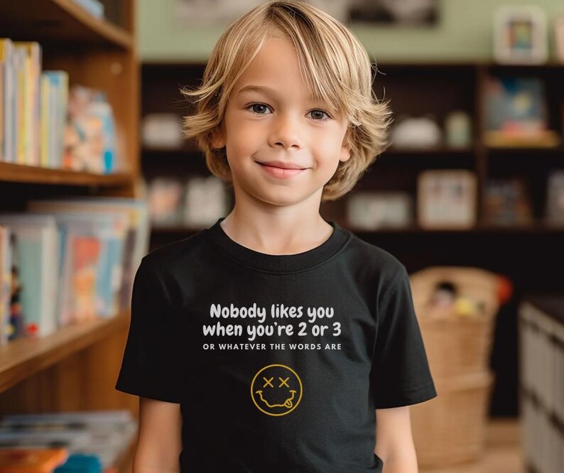 May include: A black t-shirt with white text that reads "Nobody likes you when you're 2 or 3 OR WHATEVER THE WORDS ARE" and a yellow smiley face with an X for eyes.