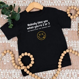 May include: Black short-sleeved t-shirt with a white graphic that says "Nobody likes you when you're 2 or 3 or whatever the words are" and a yellow smiley face with an "X" for eyes.