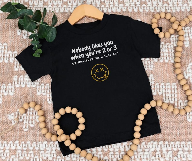 May include: Black short-sleeved t-shirt with a white graphic that says "Nobody likes you when you're 2 or 3 or whatever the words are" and a yellow smiley face with an "X" for eyes.