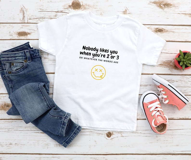 May include: White t-shirt with a yellow smiley face and the text "Nobody likes you when you're 2 or 3 OR WHATEVER THE WORDS ARE" in black.