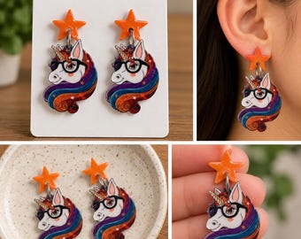 May include: A pair of dangle earrings featuring a white unicorn head with black glasses and a multi-colored rainbow mane. Each earring is topped with a small, bright orange five-pointed star charm connected by a silver-toned metal jump ring.