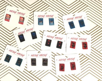 May include: A collection of Mish Mosh earrings, each pair featuring a miniature book design. The earrings are displayed on white cards with the Mish Mosh logo. The book designs vary in color and style.