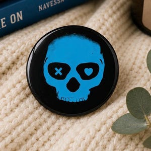 May include: A black button with a blue skull design. The skull has an X for one eye and a heart for the other. The button is round and has a glossy finish. The design is a graphic illustration.