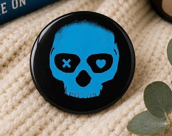 May include: A black button with a blue skull design. The skull has an X for one eye and a heart for the other. The button is round and has a glossy finish. The design is a graphic illustration.