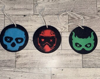 May include: Three black felt air fresheners with white strings. Each round freshener features a different graphic: a blue skull, a red helmet, and a green mask. The graphics have heart and cross details.