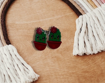 May include: Two green cactus earrings with red pots and pink flowers. The cactus has a smiling face.