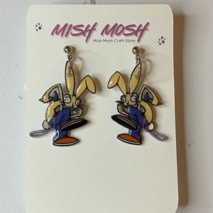 May include: A pair of clip-on earrings featuring a cartoon rabbit wearing blue overalls and holding a paintbrush. The earrings are on a white card with the text "Mish Mosh Mish Mosh Craft Store".