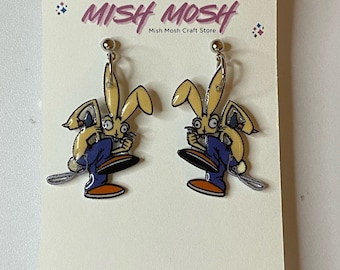 May include: A pair of clip-on earrings featuring a cartoon rabbit wearing blue overalls and holding a paintbrush. The earrings are on a white card with the text "Mish Mosh Mish Mosh Craft Store".