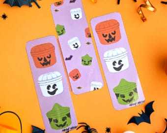 May include: Three Halloween-themed bookmarks with illustrations of jack-o'-lanterns and other spooky designs. The bookmarks are purple with orange, green, and white images. The background is orange with Halloween decorations.