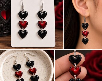 May include: Dangle earrings featuring three vertically aligned heart charms. Each earring consists of a top black heart, a middle metallic red heart, and a bottom black heart, all connected by silver-toned metal jump rings with hook-style ear wires.