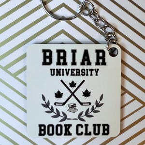 May include: White square keychain with black text that reads "Briar University Book Club". The keychain features a graphic of two crossed hockey sticks with maple leaves and a stack of books.