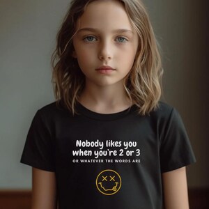 May include: Black t-shirt with white text that reads "Nobody likes you when you're 2 or 3 or whatever the words are" and a yellow smiley face with an X for eyes.