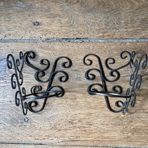 Op de afbeelding: Twee zwarte smeedijzeren wandkandelaars met een krullend ontwerp. Elke kandelaar heeft een cirkelvormige houder en decoratieve krullen. De kandelaars worden op een houten oppervlak gepresenteerd.