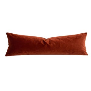 May include: A long, rectangular lumbar pillow featuring a solid, deep rust-orange velvet fabric. The pillow has a smooth, soft-textured surface with subtle light reflections across the material, creating a consistent color tone throughout the item.