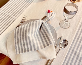 May include: A white and gray striped napkin is folded and placed on a white and gray striped tablecloth. The napkin is held in place with a silver ring. A silver fork, knife, and spoon are placed on the table next to the napkin. A glass of water is on the table to the right of the napkin.