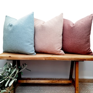 May include: Three decorative throw pillows in different colors: light blue, light pink, and dusty rose. The pillows are arranged on a wooden bench.