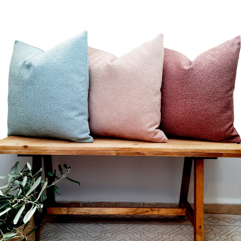May include: Three decorative throw pillows in different colors: light blue, light pink, and dusty rose. The pillows are arranged on a wooden bench.