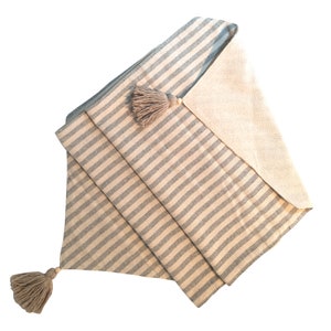 Puede incluir: Camino de mesa plegado con un estampado de rayas grises y beige. El camino de mesa tiene dos borlas beige con cuentas de madera en cada extremo. La tela está doblada para mostrar el reverso, que es de color beige liso.