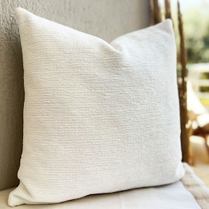 May include: A white, textured throw pillow with a subtle woven pattern. The pillow is shown on a light beige surface.