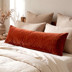 May include: A long, rectangular bolster pillow in a solid burnt orange velvet fabric. The pillow is positioned horizontally across the front of a bed, layered in front of several neutral-toned beige and cream pillows on a white linen bedspread.