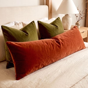 May include: A long rectangular rust-colored velvet lumbar pillow positioned in front of two square olive green velvet throw pillows. The pillows are arranged on a bed with light-colored bedding, featuring a smooth, soft-touch fabric texture.