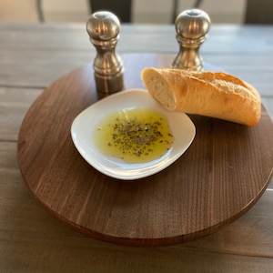 May include: A wooden serving tray with a white dish filled with olive oil and herbs, a baguette, and two silver salt and pepper shakers.