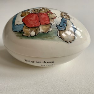 Può includere: Scatola portaoggetti in ceramica bianca a forma di uovo con un'illustrazione di Peter Rabbit. La scatola presenta una scena con Peter Rabbit e altri tre conigli. Il testo "Peter sat down to rest" è stampato sul fondo della scatola.