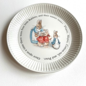 Op de afbeelding: Een wit keramiek bord met een gegolfde rand heeft een vintage illustratie van vier konijnen. De tekst luidt "Once upon a time there were four little Rabbits, and their names were- Flopsy, Mopsy, Cotton-tail, and Peter."