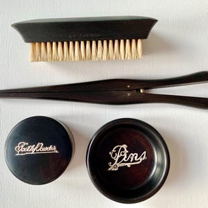 May include: A vintage grooming set featuring a dark wood brush, a tweezer, and two small containers. One container is labeled "Tooth Powder" and the other "Pins" in elegant script. The set is arranged on a white surface.