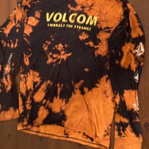 May include: A black and orange tie-dye long-sleeve shirt with the text "Volcom Embrace the Strange" in yellow.
