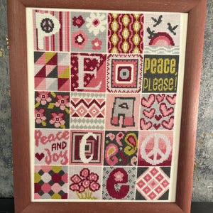 Könnte beinhalten: Ein gerahmtes Kreuzstich-Kunstwerk mit einem braunen Holzrahmen. Das Kunstwerk zeigt ein Raster aus farbenfrohen Quadraten mit verschiedenen Designs, darunter Blumen, Herzen und die Worte "peace please" und "peace and joy". Die Farbpalette umfasst Rosa, Rot und Grau.