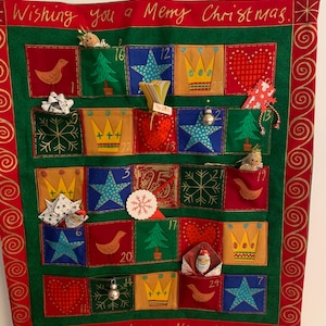 Könnte beinhalten: Ein grüner Filz-Adventskalender mit roten, gelben und blauen Taschen. Jede Tasche hat ein anderes Design, darunter Sterne, Kronen, Herzen und Vögel. Der Kalender ist mit dem Text "Wishing you a Merry Christmas" und "and a happy New Year" verziert.