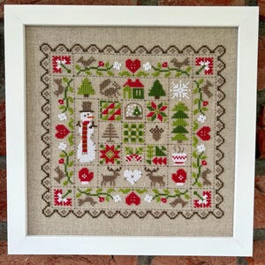 Könnte beinhalten: Ein gerahmtes Kreuzstichbild mit winterlichem Feiertagsmotiv. Das Design zeigt einen Schneemann, Bäume, Herzen, Fäustlinge und Rentiere, alles in Rot, Grün und Weiß. Das Kunstwerk ist in einem weißen quadratischen Rahmen gerahmt.