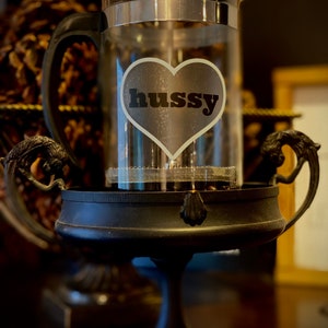 Könnte beinhalten: Ein schwarzer Metallständer mit einer gläsernen French Press Kaffeemaschine. Die Kaffeemaschine hat einen herzförmigen Aufkleber mit dem Wort "hussy" darauf.