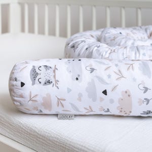 Op de afbeelding: Een wit en grijs baby nest met een bedrukt patroon van uilen, wolken en bladeren. Het nest is gemaakt van zachte stof en is ontworpen om een veilige en comfortabele slaapruimte voor een baby te bieden. Het nest is gelabeld "Loolay".