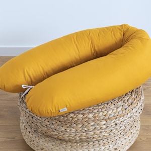 Op de afbeelding: Mosterdgeel U-vormig lichaamskussen met witte banden, rustend op een geweven rieten mand. Het kussen is gemaakt van zachte stof en is ontworpen voor comfort en ondersteuning. Het kussen heeft een warme, uitnodigende kleur.