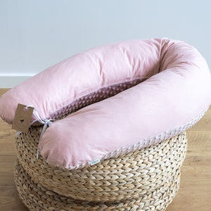 Könnte beinhalten: Ein hellrosa, U-förmiges Schwangerschaftskissen mit weicher, samtiger Textur. Das Kissen liegt auf einem geflochtenen Weidenkorb. Das Kissen hat ein Etikett mit einer Schnur befestigt. Das Kissen ist für Komfort und Unterstützung während der Schwangerschaft konzipiert.