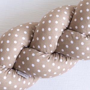Op de afbeelding: Gevlochten babybumper in een neutrale bruintint met witte stippen. De bumper is gemaakt van zachte stof en is ontworpen om de baby te beschermen tegen stoten en tocht. De merknaam "LOOLAY" is zichtbaar op een klein label.